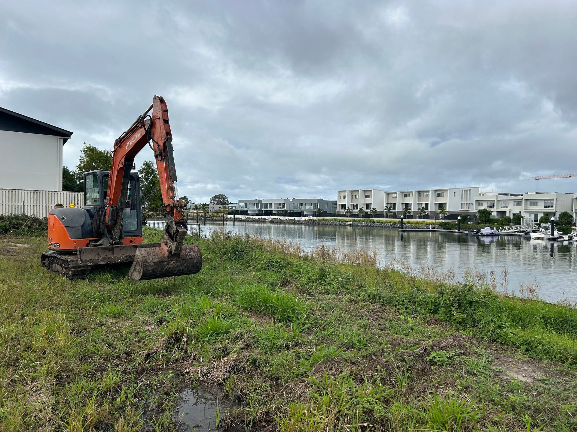 Orange excavator on a grassy riverbank beside calm water and modern houses under cloudy skies