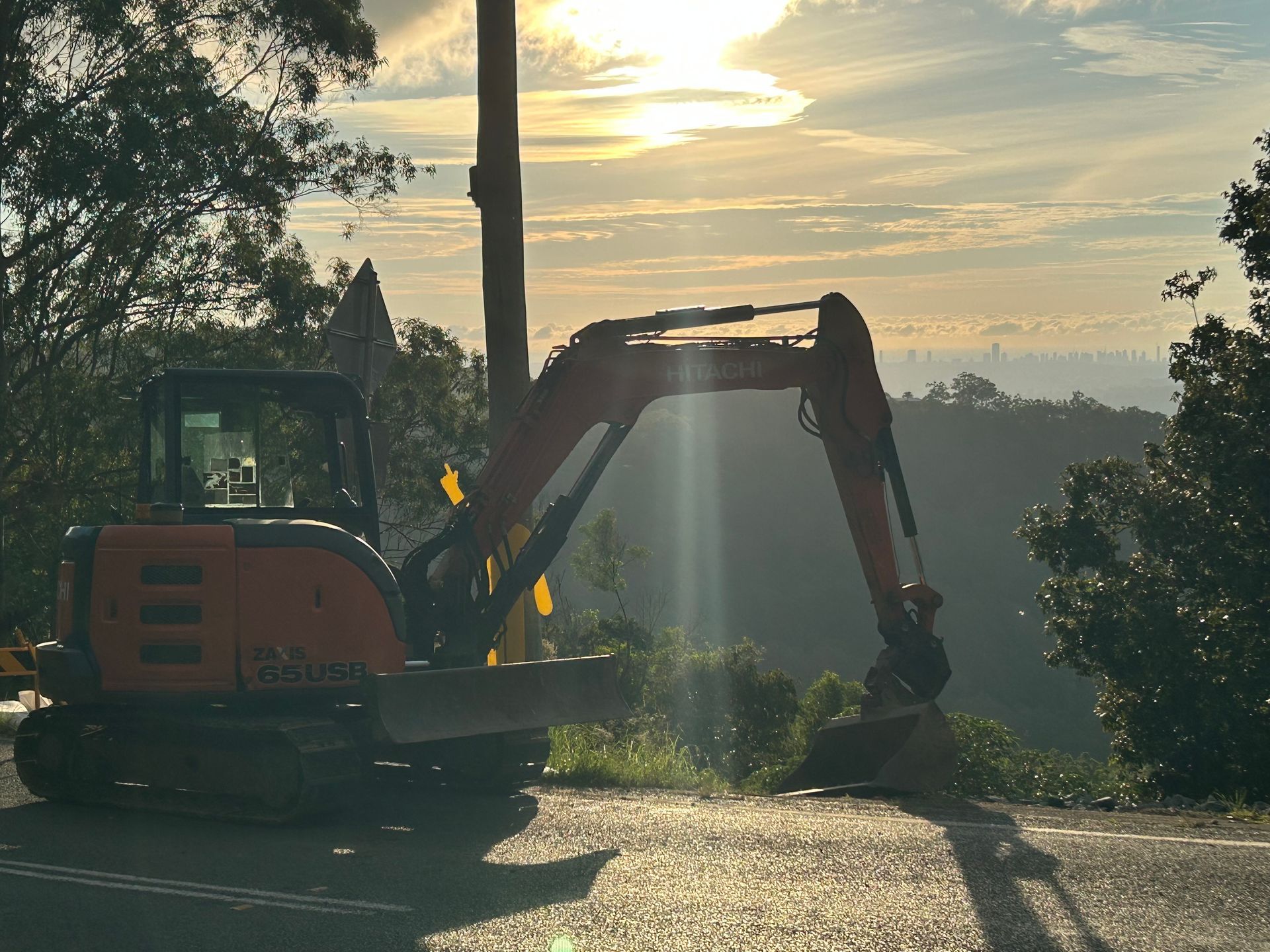 Excavator silhouetted at sunset on a roadside, working beside trees and hills