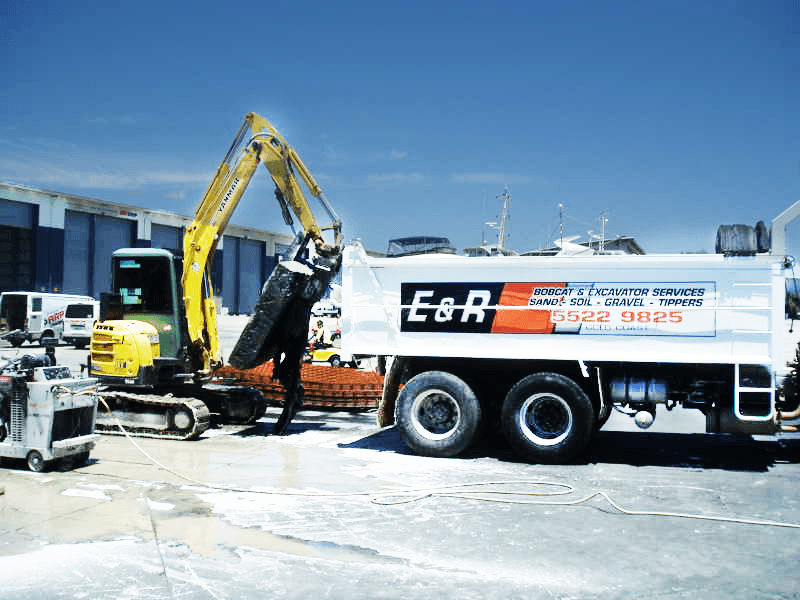 Yellow excavator loading debris into white E&R dump truck at a dusty worksite under a blue sky