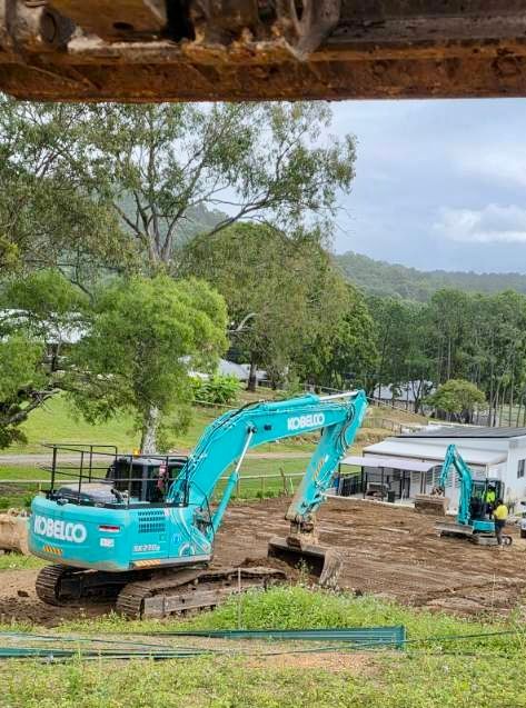 Turquoise excavators at a construction site on a grassy hillside with trees in the background