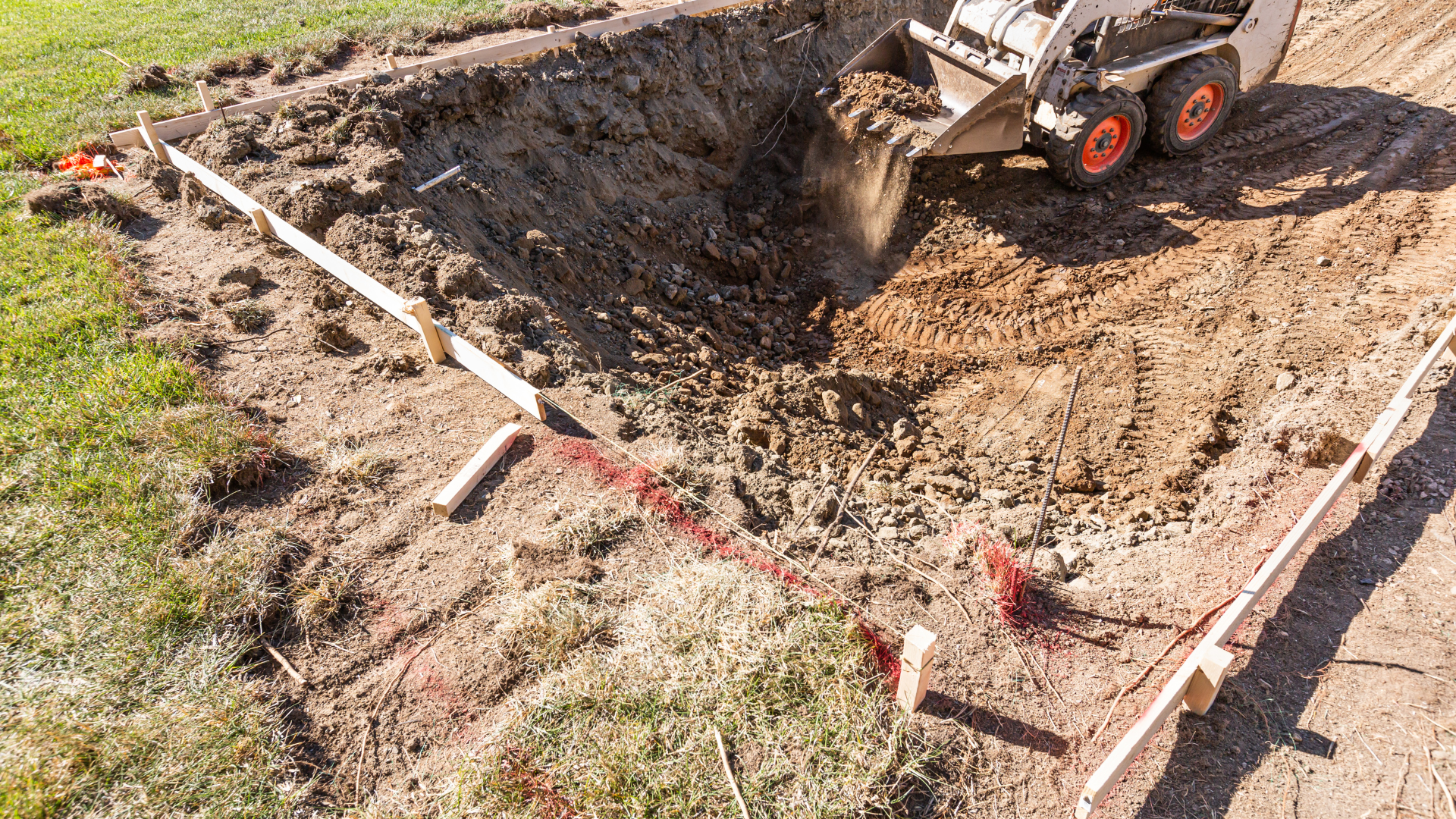 Small excavator digging a large dirt pit beside a marked construction area