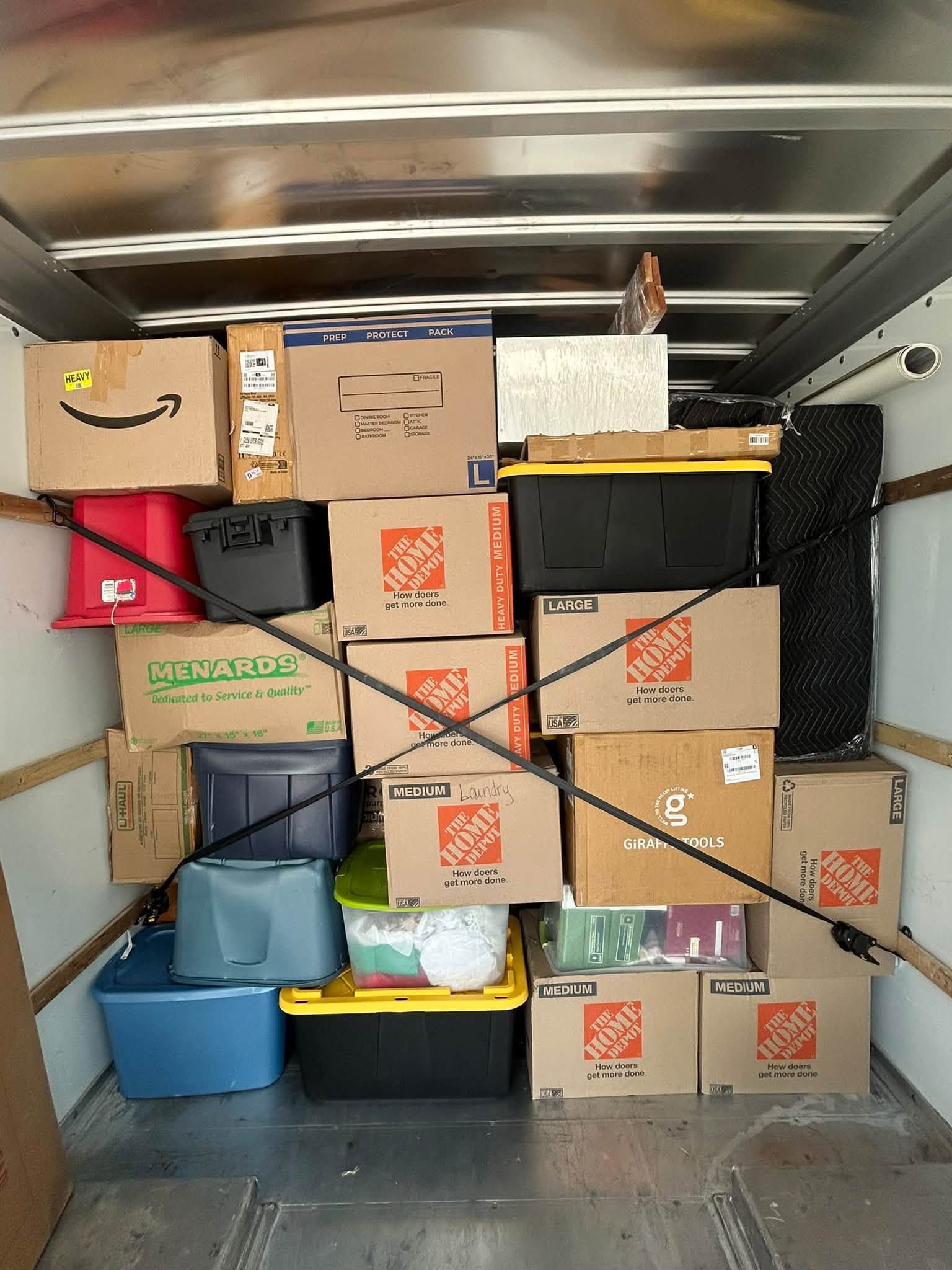 Packed moving truck with stacked cardboard boxes, bins, and a wrapped item secured inside the cargo area.