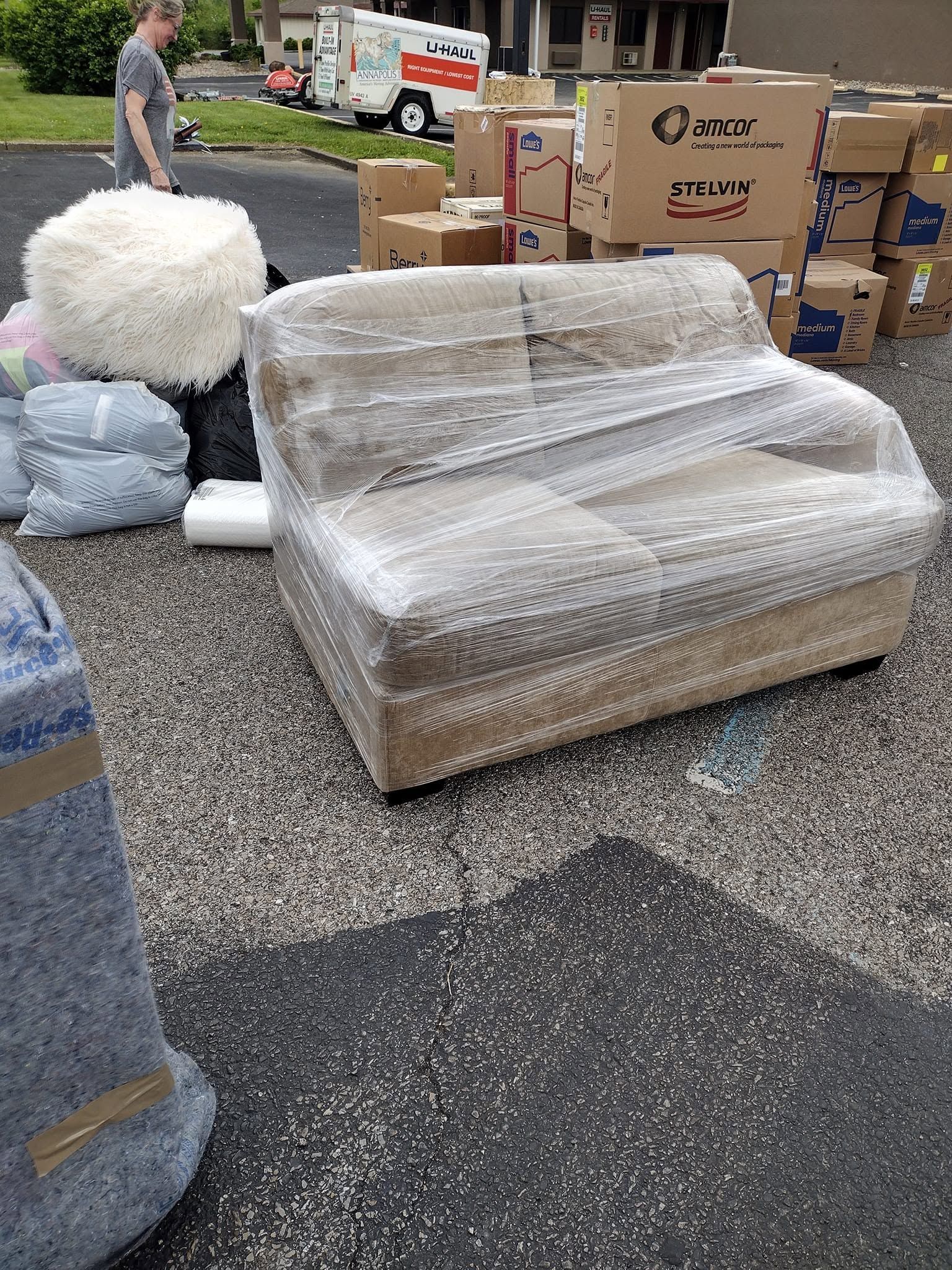 Shrink-wrapped couch and boxes on a gravel lot, with a worker and moving truck in the background.