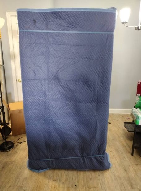 Blue padded moving blanket covering a tall object in a room with wood flooring and a lamp nearby
