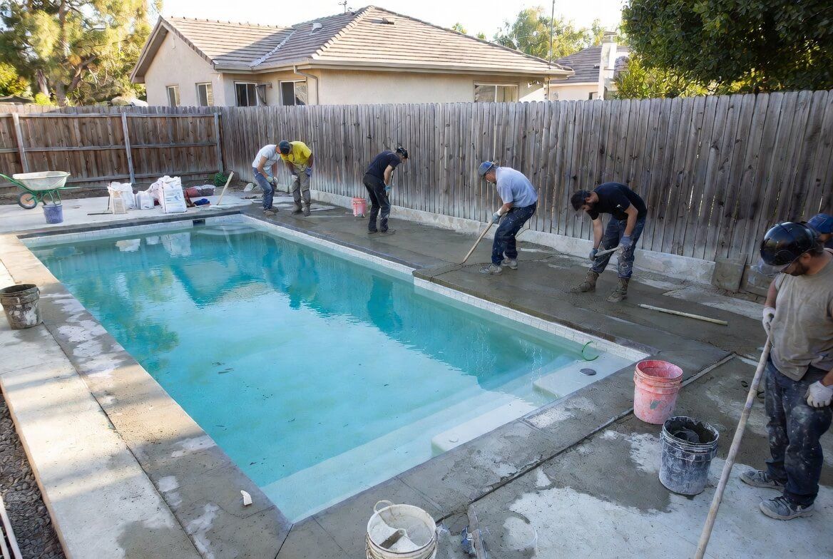 A gloved hand uses a trowel to smooth wet concrete along a brick border outdoors.