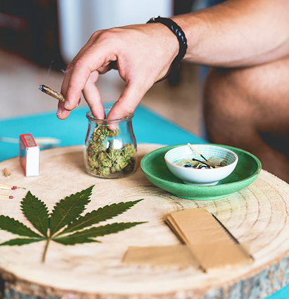 Hand reaching into a small glass jar of cannabis beside a green bowl and cannabis leaf on a wooden table