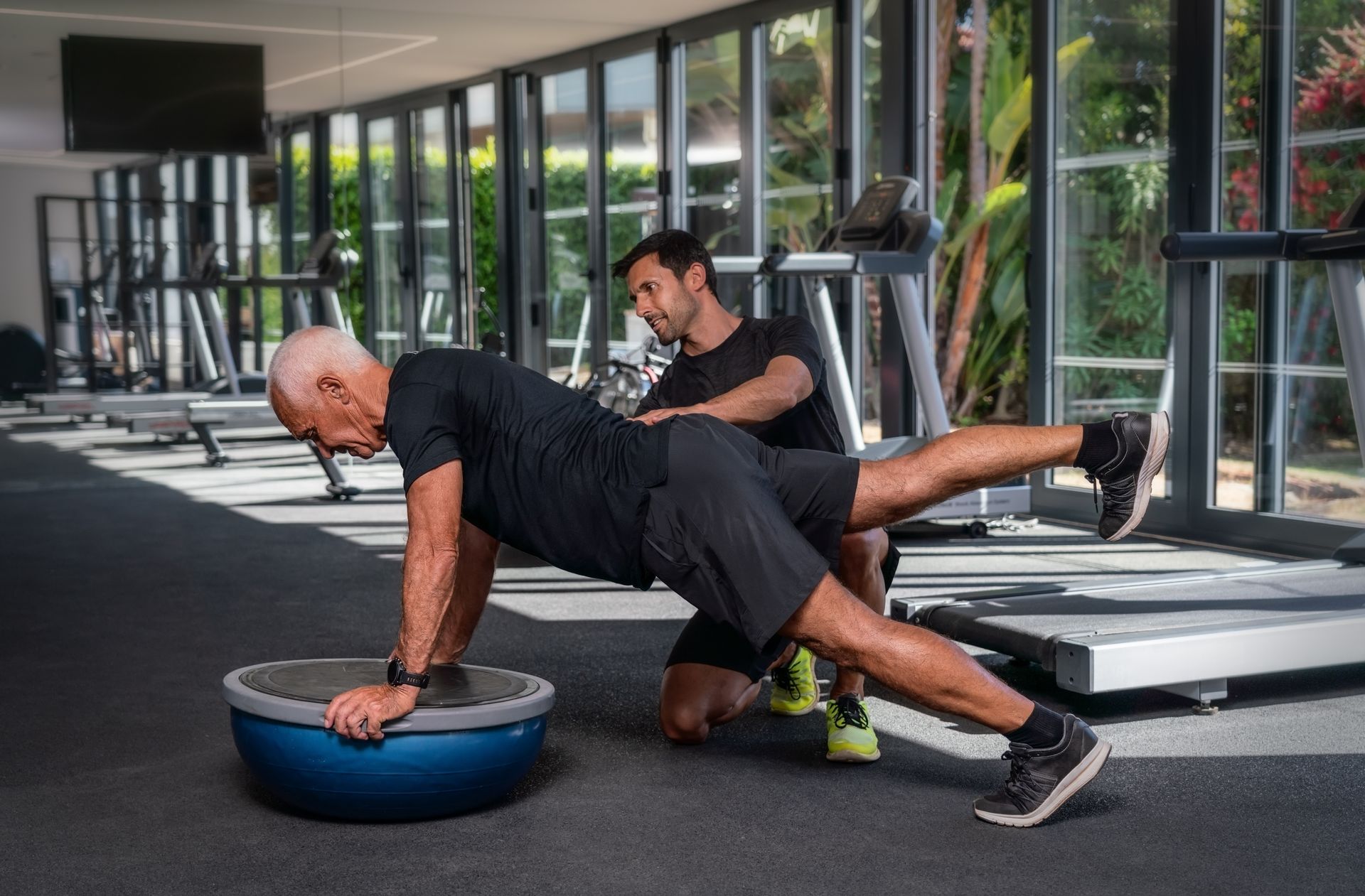 Two people doing a plank exercise in a gym, one on a balance dome and one spotting behind him. Two people doing a plank exercise in a gym, one on a balance dome and one spotting behind him.