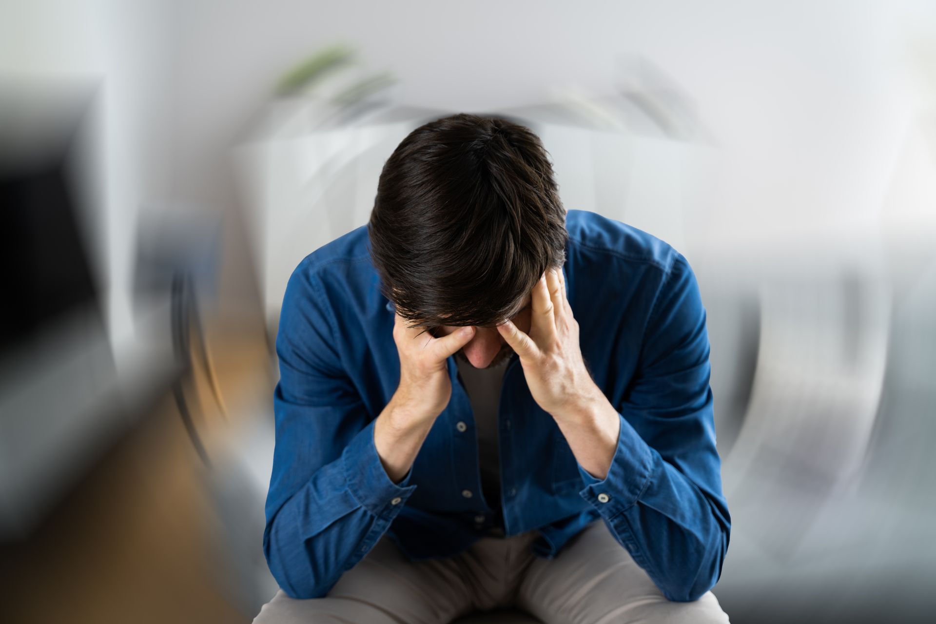 Man sitting with head in hands, appearing stressed, in a blurred indoor setting