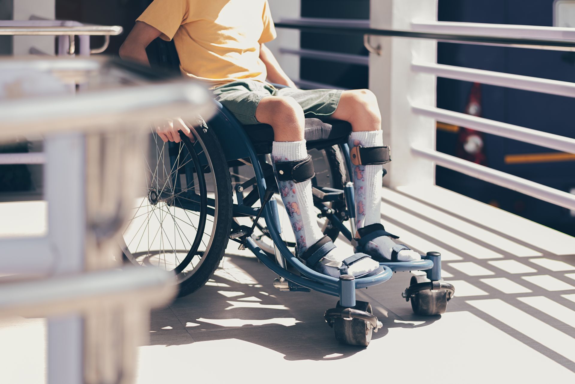 Person in a wheelchair on a sunlit balcony, wearing a yellow shirt and black leg braces.