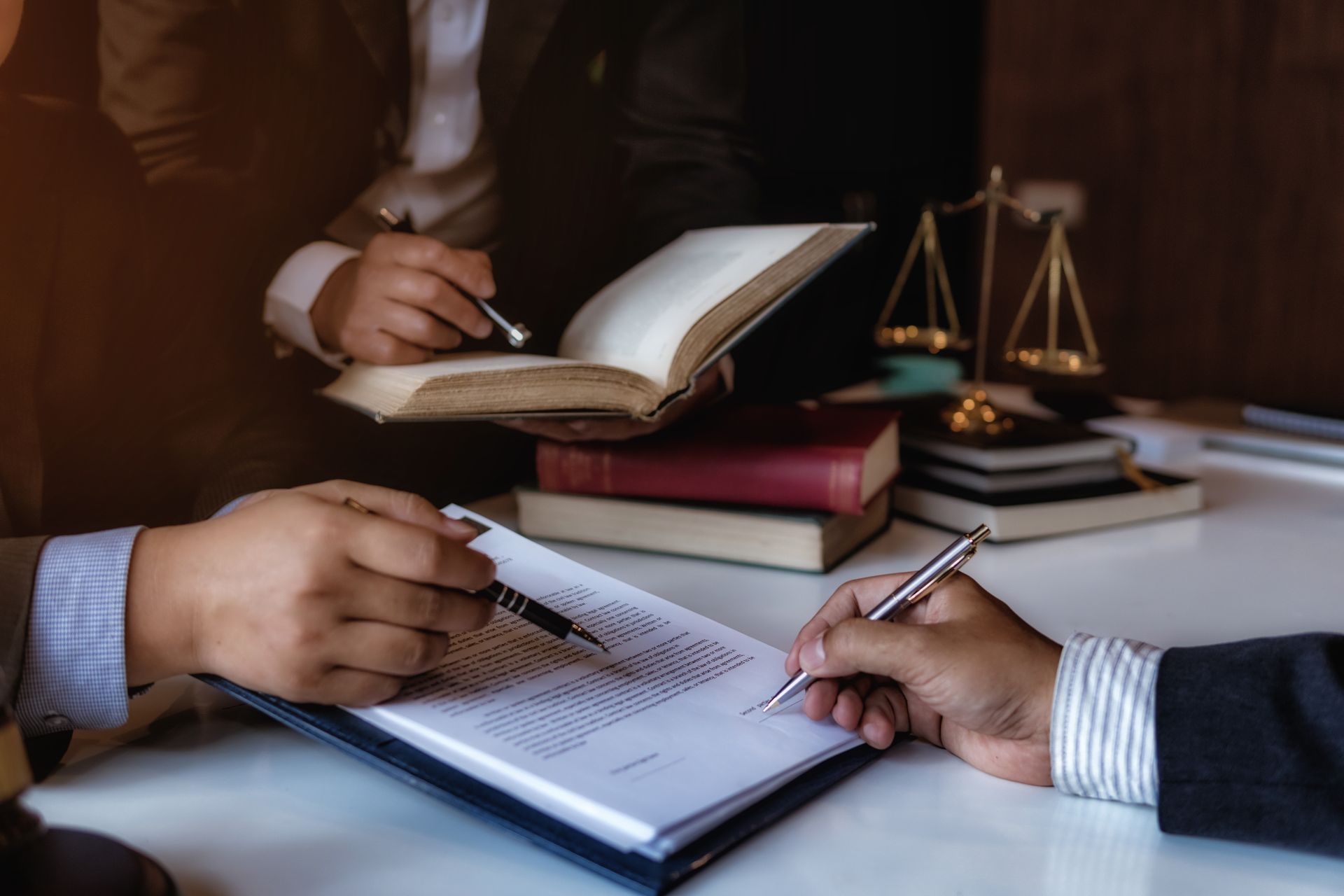 Hands signing a document at a desk with an open notebook, books, and a justice scale nearby