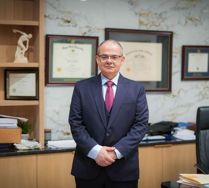 Man in a navy suit and pink tie standing in an office with framed certificates on the wall