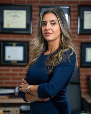 Portrait of a woman standing with folded arms in an office, with framed certificates on a brick wall behind her.
