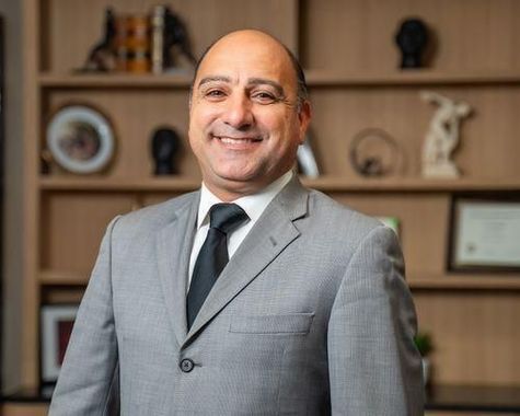 Smiling man in a gray suit standing in an office with shelves and framed decor behind him