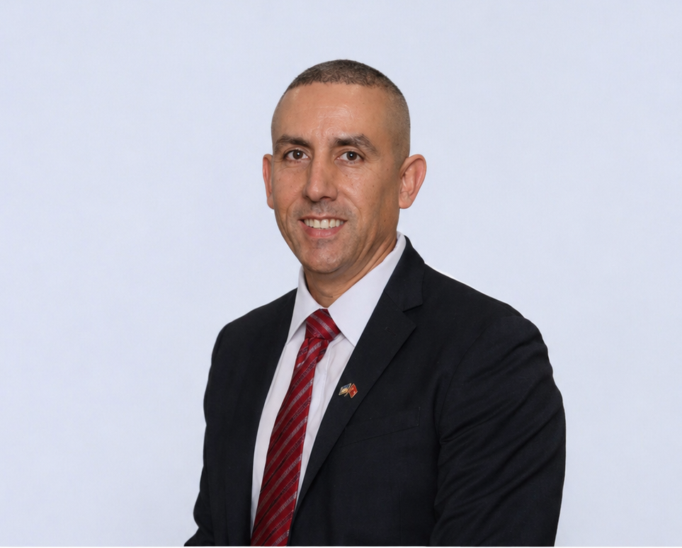 Man in a dark suit with a red striped tie, standing indoors and smiling toward the camera