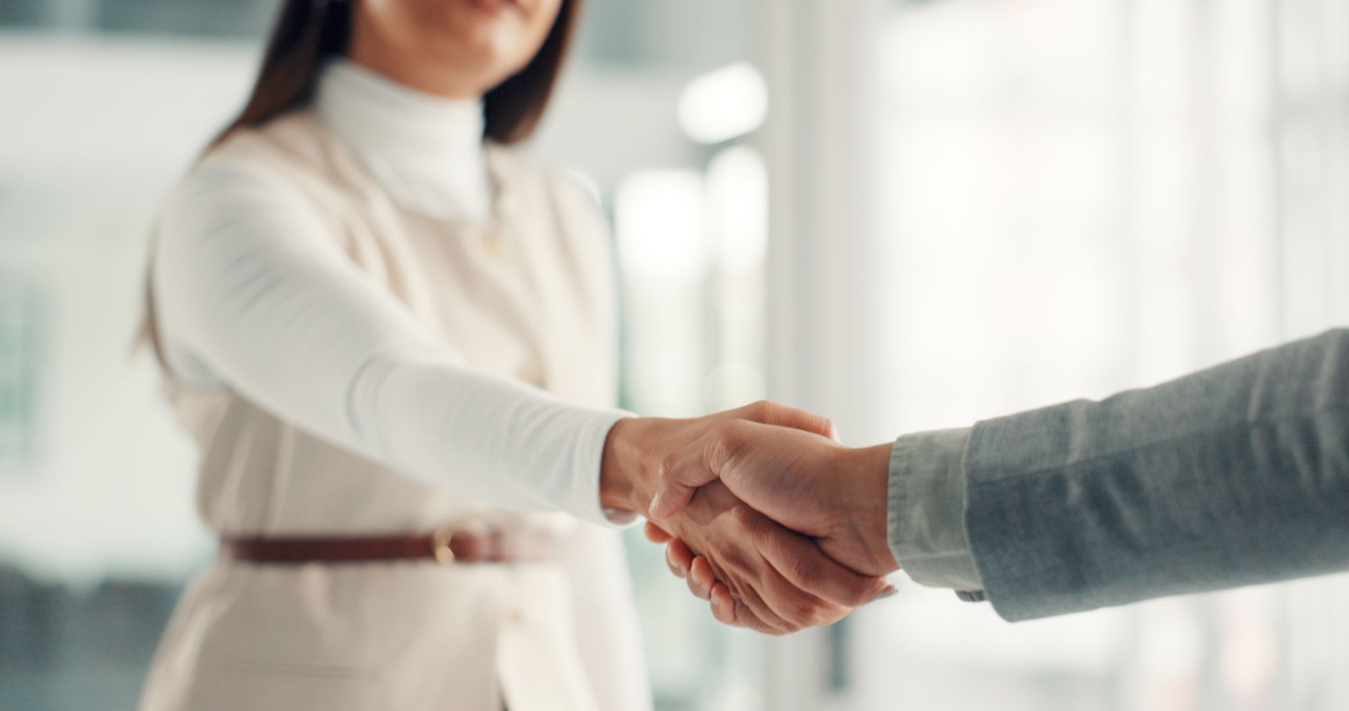Two people shaking hands in a bright office, with one wearing a white top and the other a gray sleeve