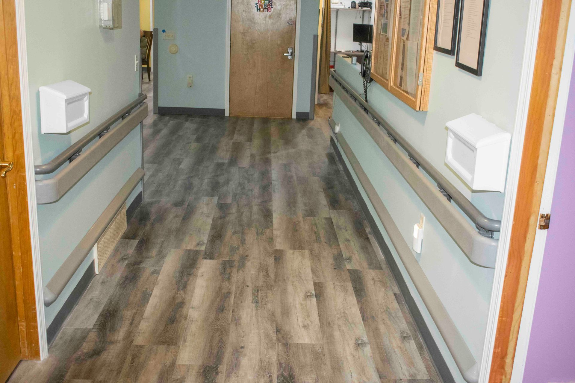 Empty hospital hallway with wood-look flooring, pale walls, handrails, and doors at the end.