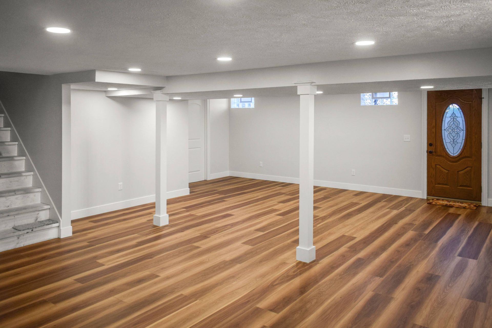 Empty finished basement with wood floors, white walls and columns, recessed lights, and stairs on the left.