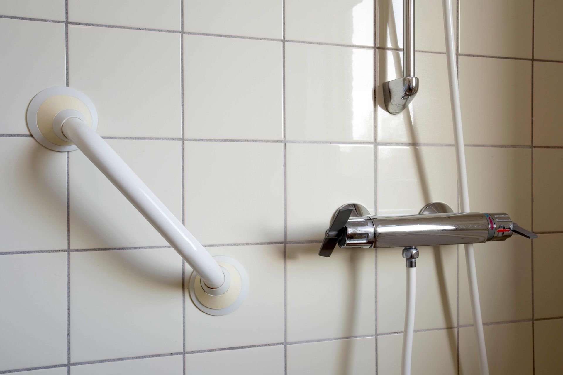 Bathroom shower with a white grab bar, tiled wall, and chrome shower controls with handheld hose.