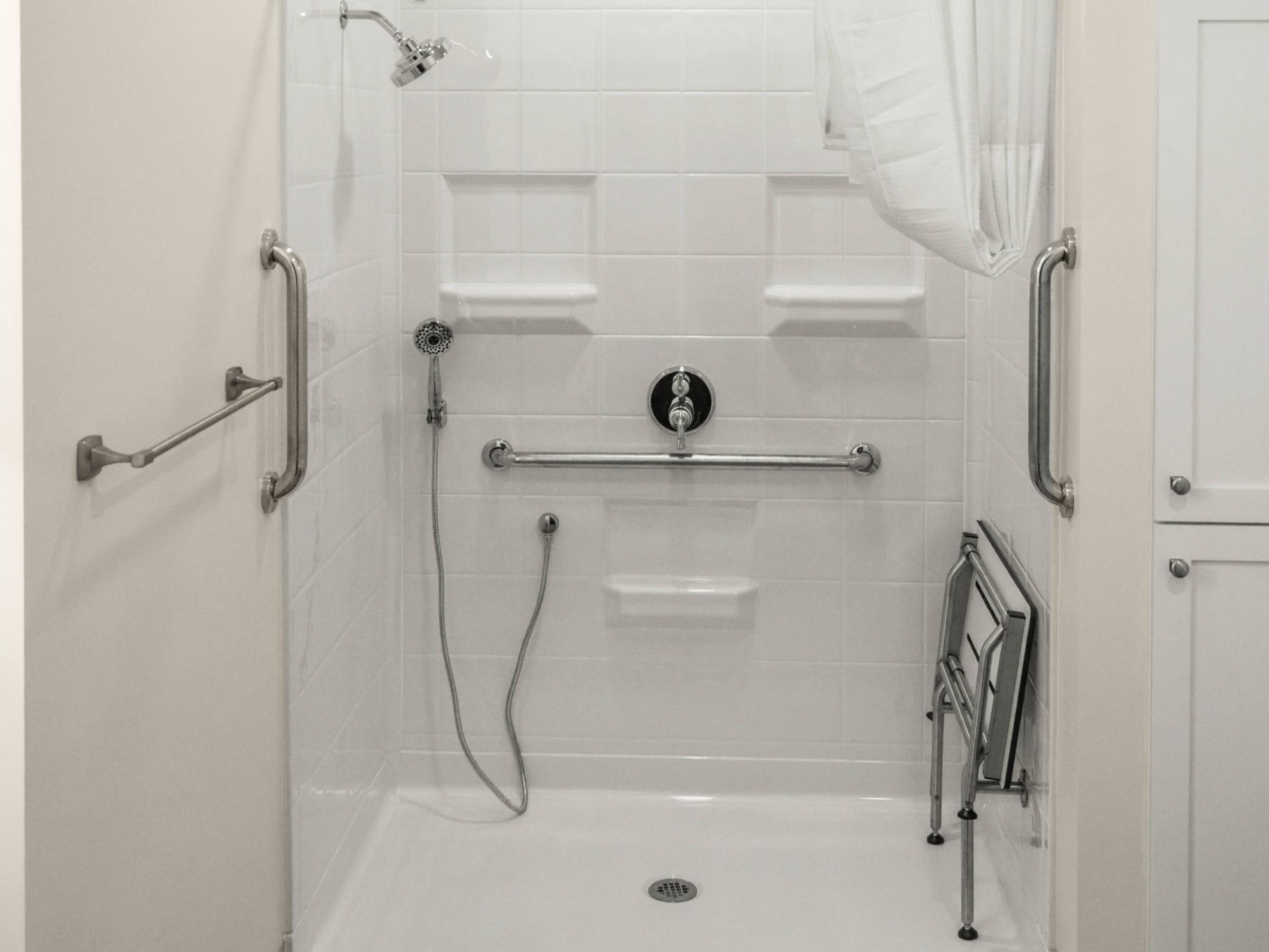 Accessible shower with grab bars, shower head, folding seat, and white walls.