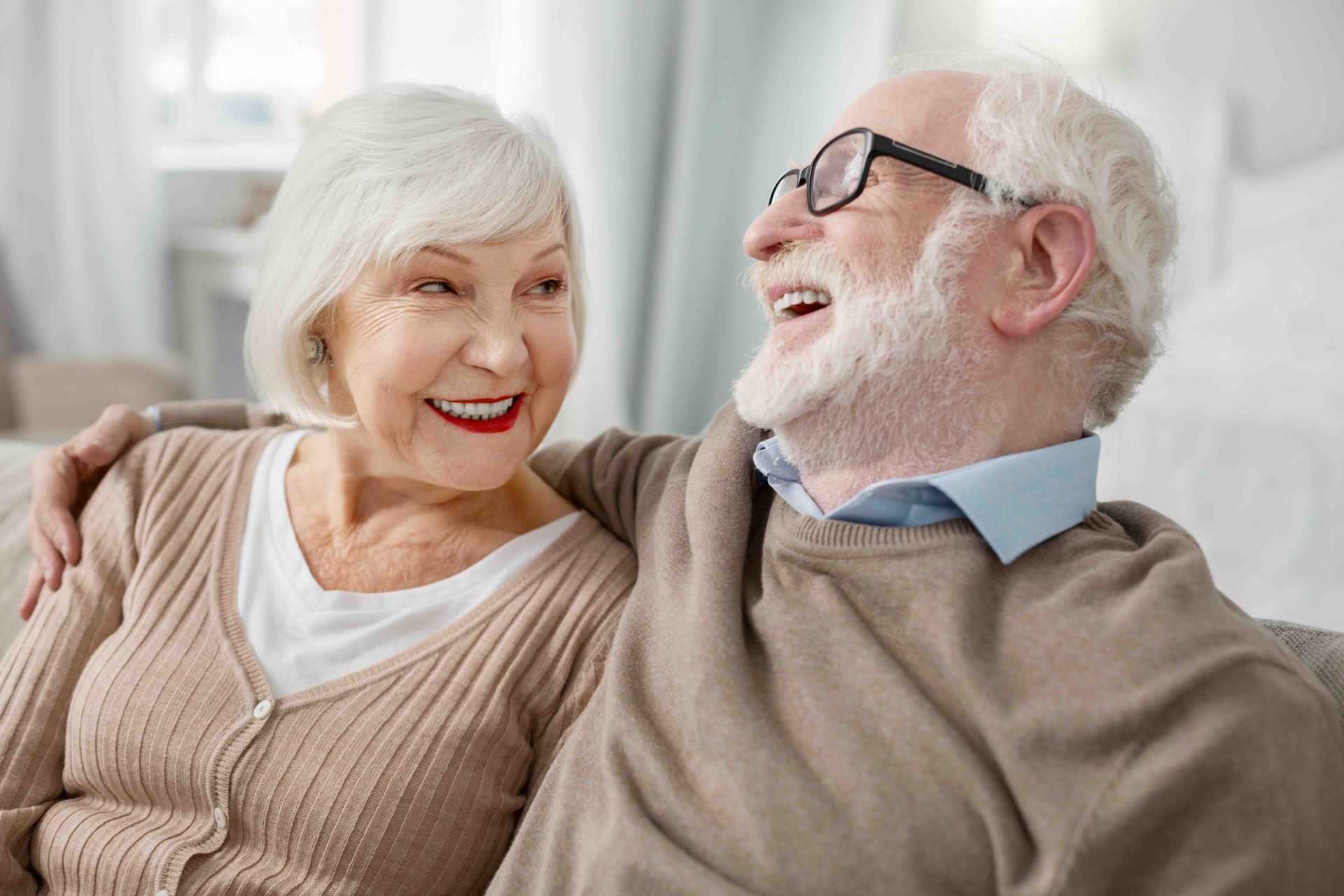 A smiling couple embraces while sitting indoors in a light-filled room.