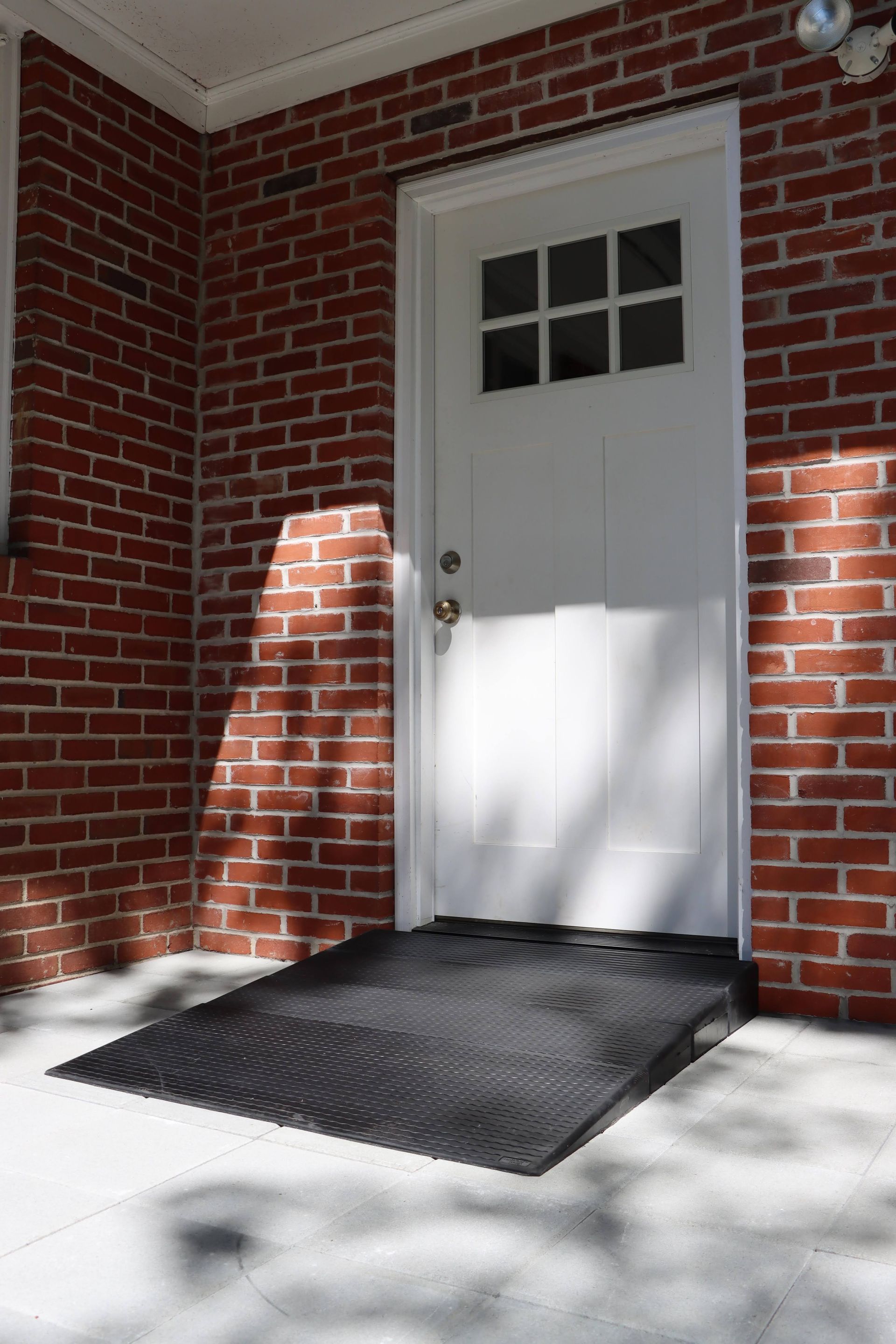White door with window panes in a red brick wall, with a black ramp mat at the entrance.