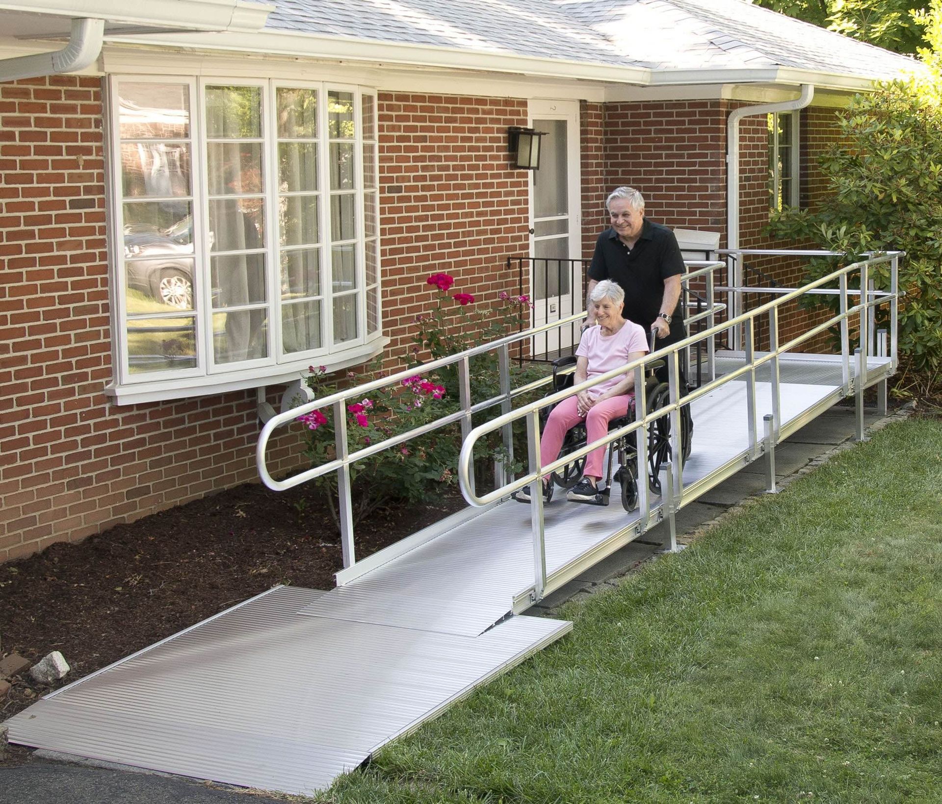 Person in a wheelchair on a ramp outside a brick house, assisted by another person behind them