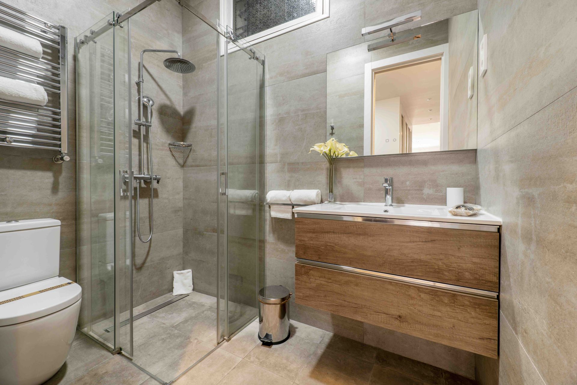 Modern bathroom with glass shower, wooden vanity, and toilet. Gray tile walls.