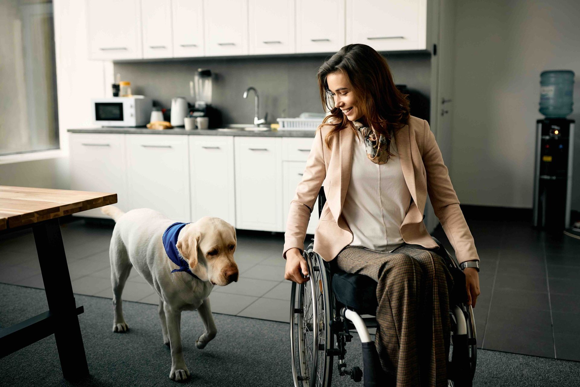 Woman in wheelchair smiles at a Labrador guide dog in a kitchen.