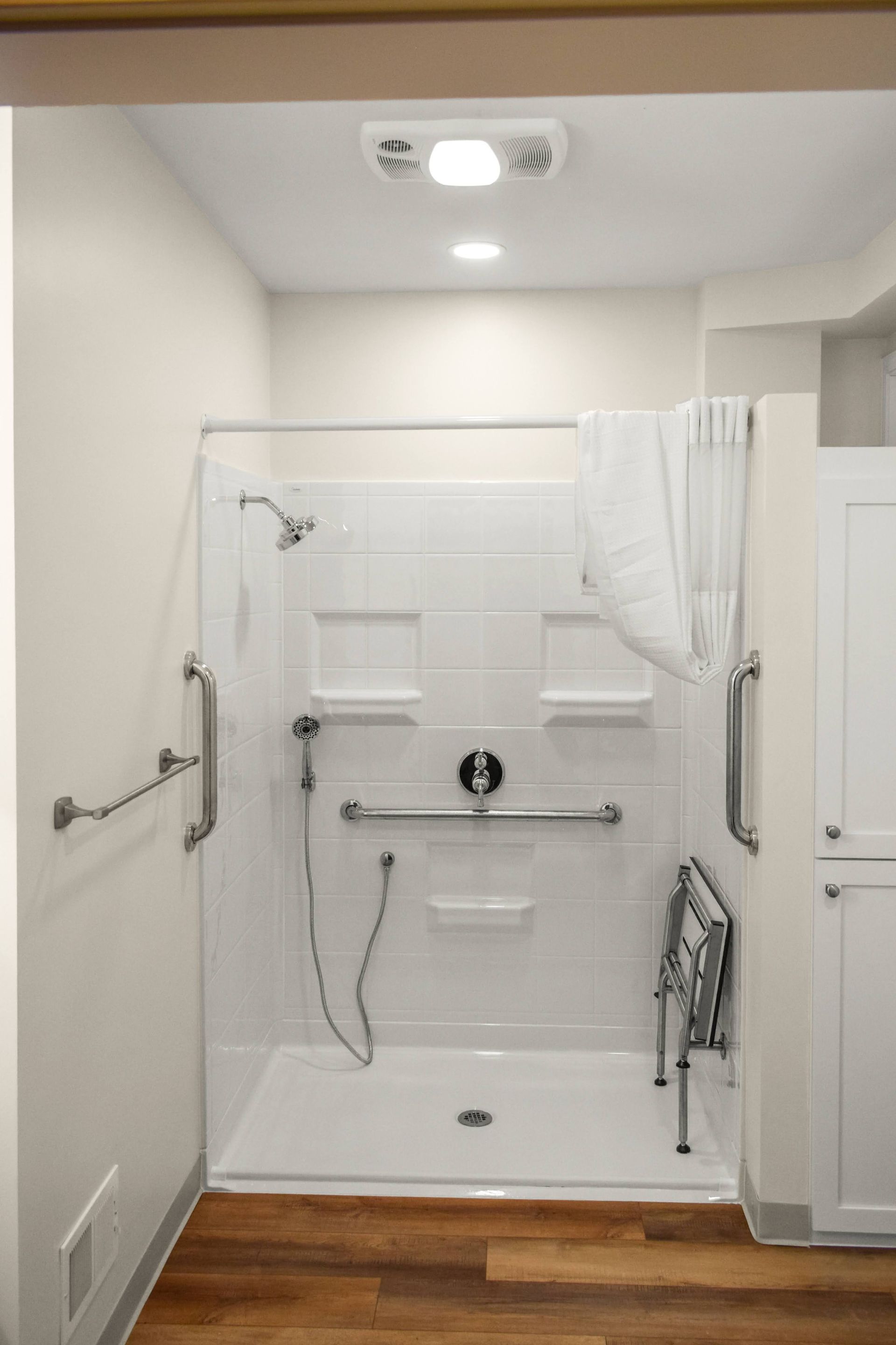 Accessible bathroom with grab bars, shower seat, and white tiled walls.