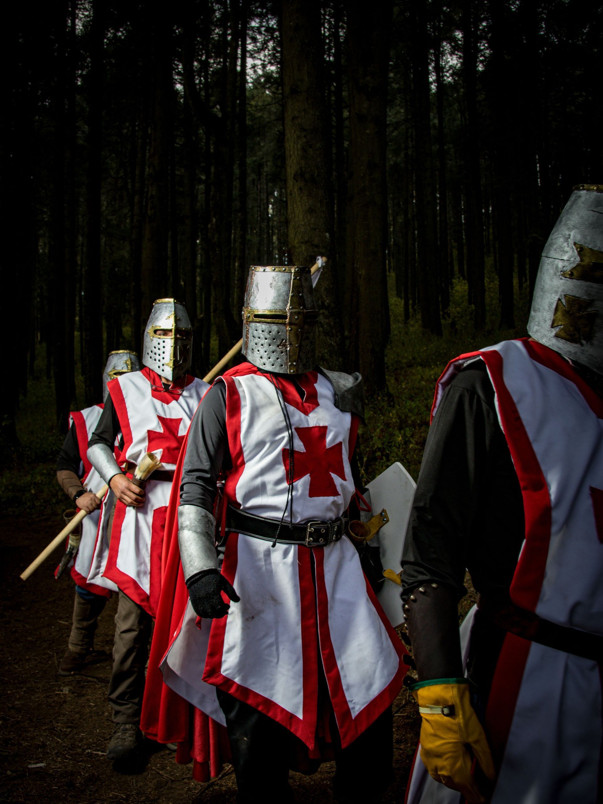Knights in white robes with red crosses stand in a dark forest, one holding a sword.