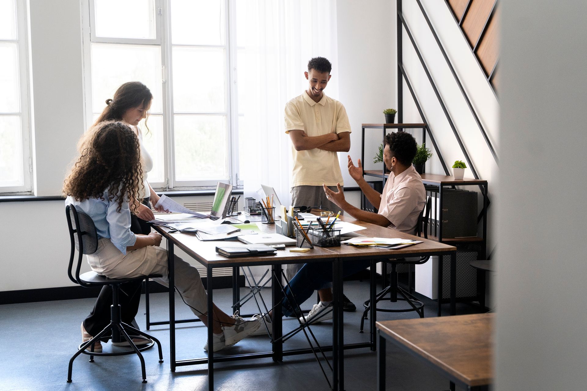 Four people collaborating at a desk in an office. One man gestures while talking, others listen.