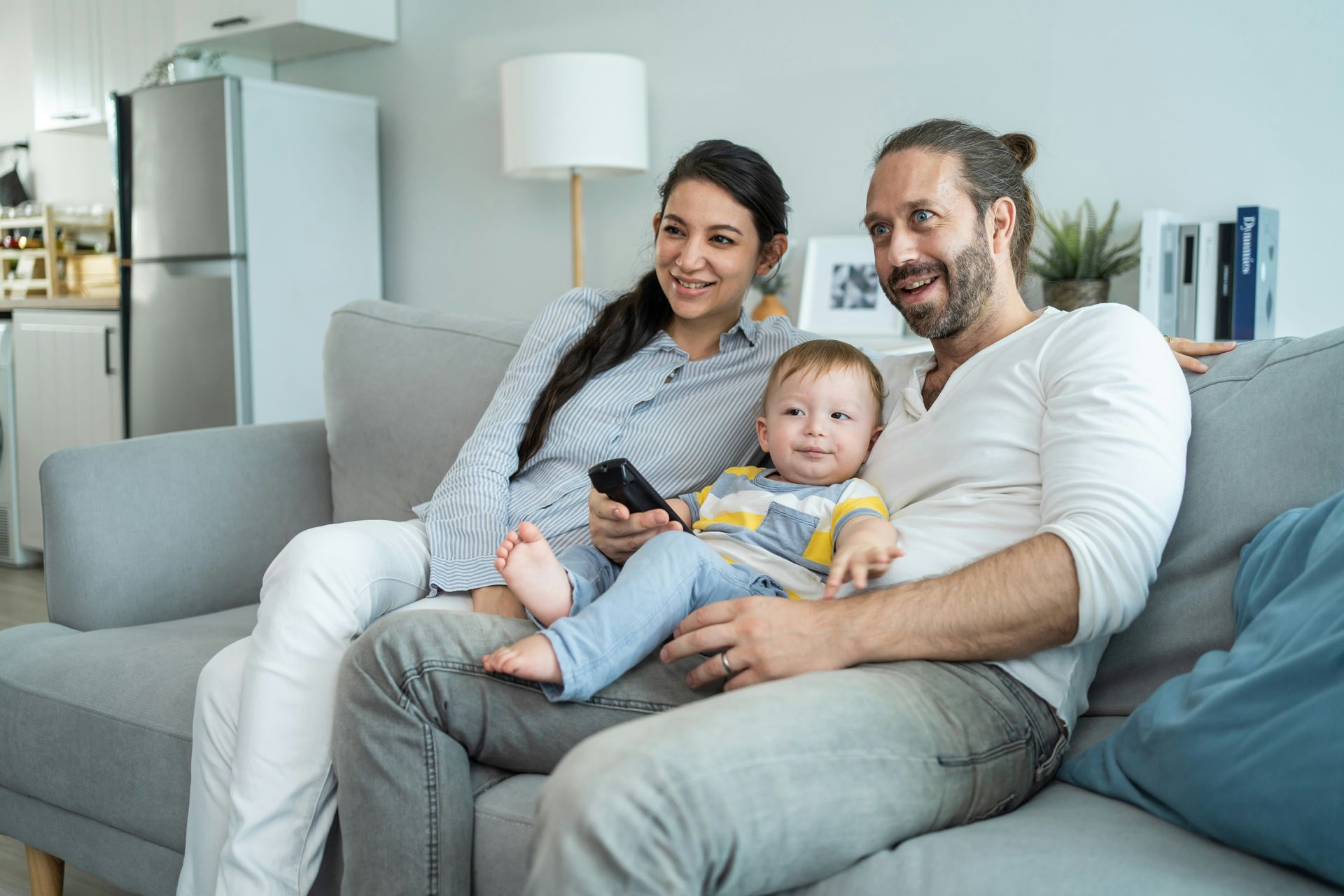 A family sits on a couch, watching TV. A baby sits on the father's lap, smiling. The mother holds a remote.