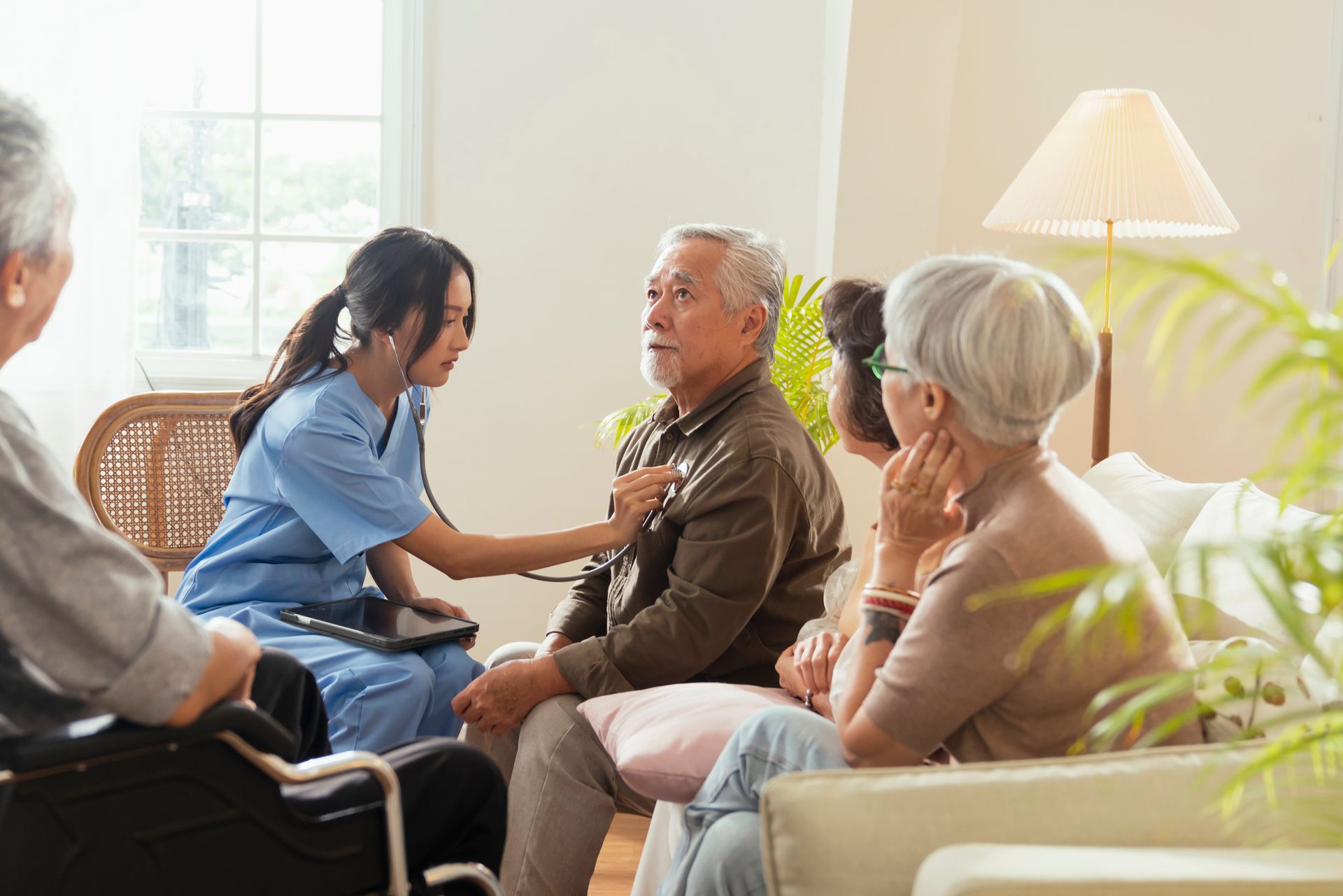 Nurse using a stethoscope on a man in a living room, other seniors seated nearby.