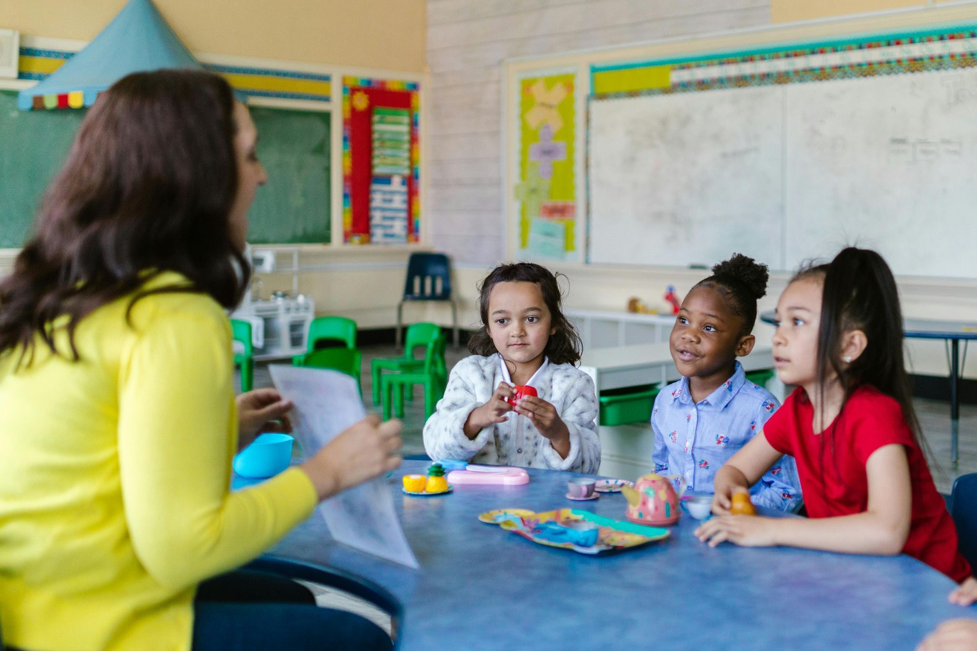 Teacher with papers speaks to children at a blue table in a classroom.