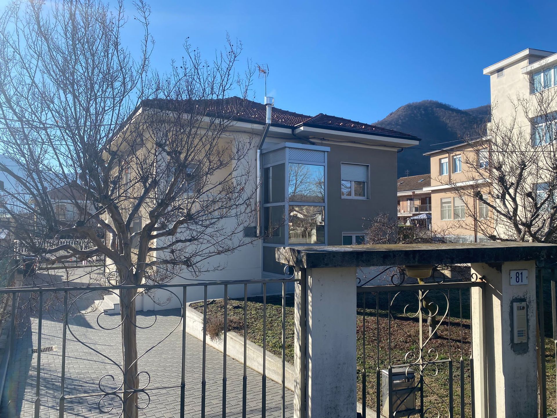 Casa dietro una recinzione in ferro battuto con alberi spogli, giornata di sole, montagne sullo sfondo.