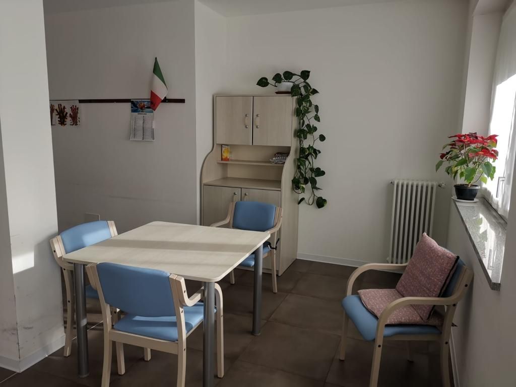 Una piccola zona pranzo con tavolo, sedie, mobiletto, piante e termosifone. Alla parete è appesa una bandiera italiana.