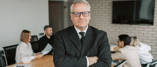 Confident man in a suit stands in a conference room with coworkers blurred in the background
