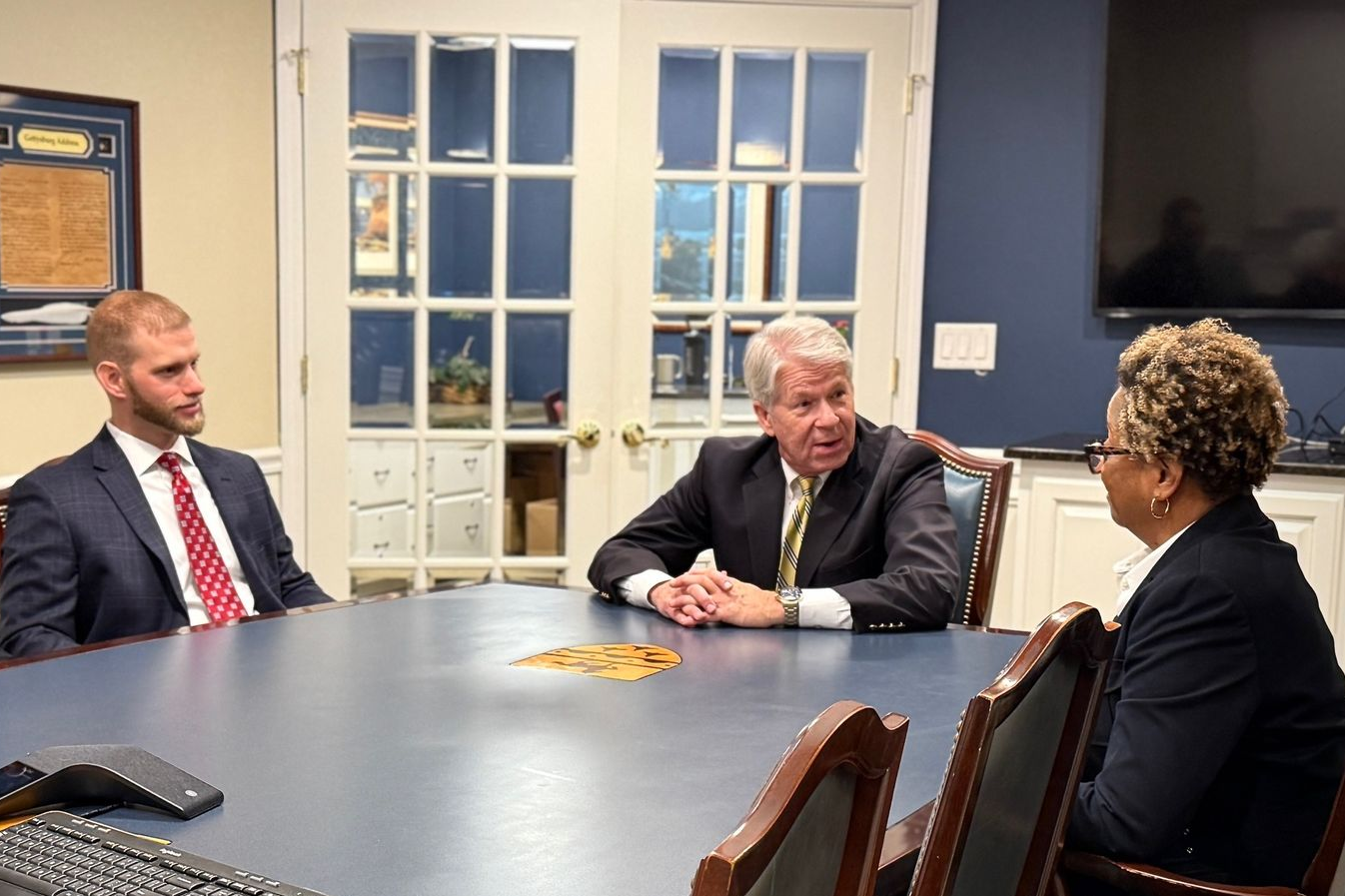 Three people in a conference room discuss a meeting around a dark table.