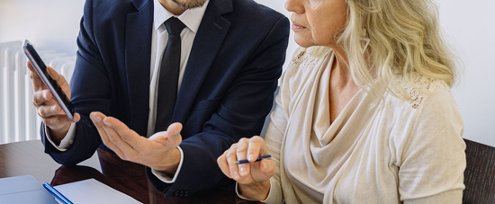 Two businesspeople discussing a document at a desk, one holding a tablet and the other gesturing.