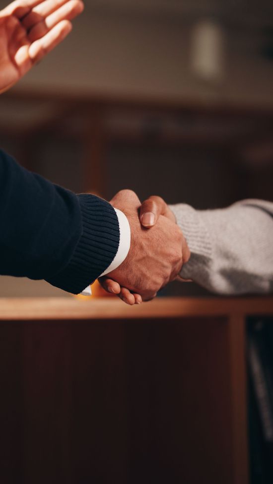 Two people shaking hands across a table, one in a dark sweater and one in a light sweater.