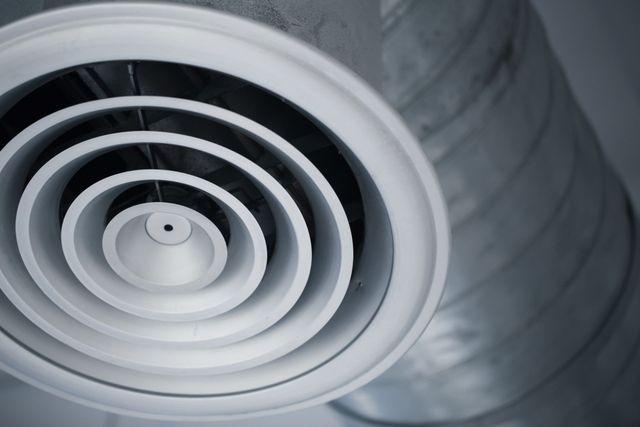 A white, circular HVAC diffuser with concentric rings mounted to a metallic industrial duct.