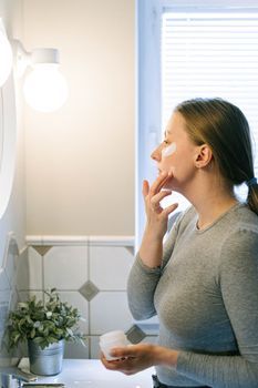 Young woman applying moisturizer to her cheek