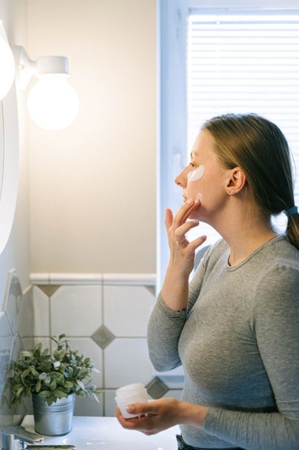 Woman applying moisturizer at home in front of a bathroom mirror