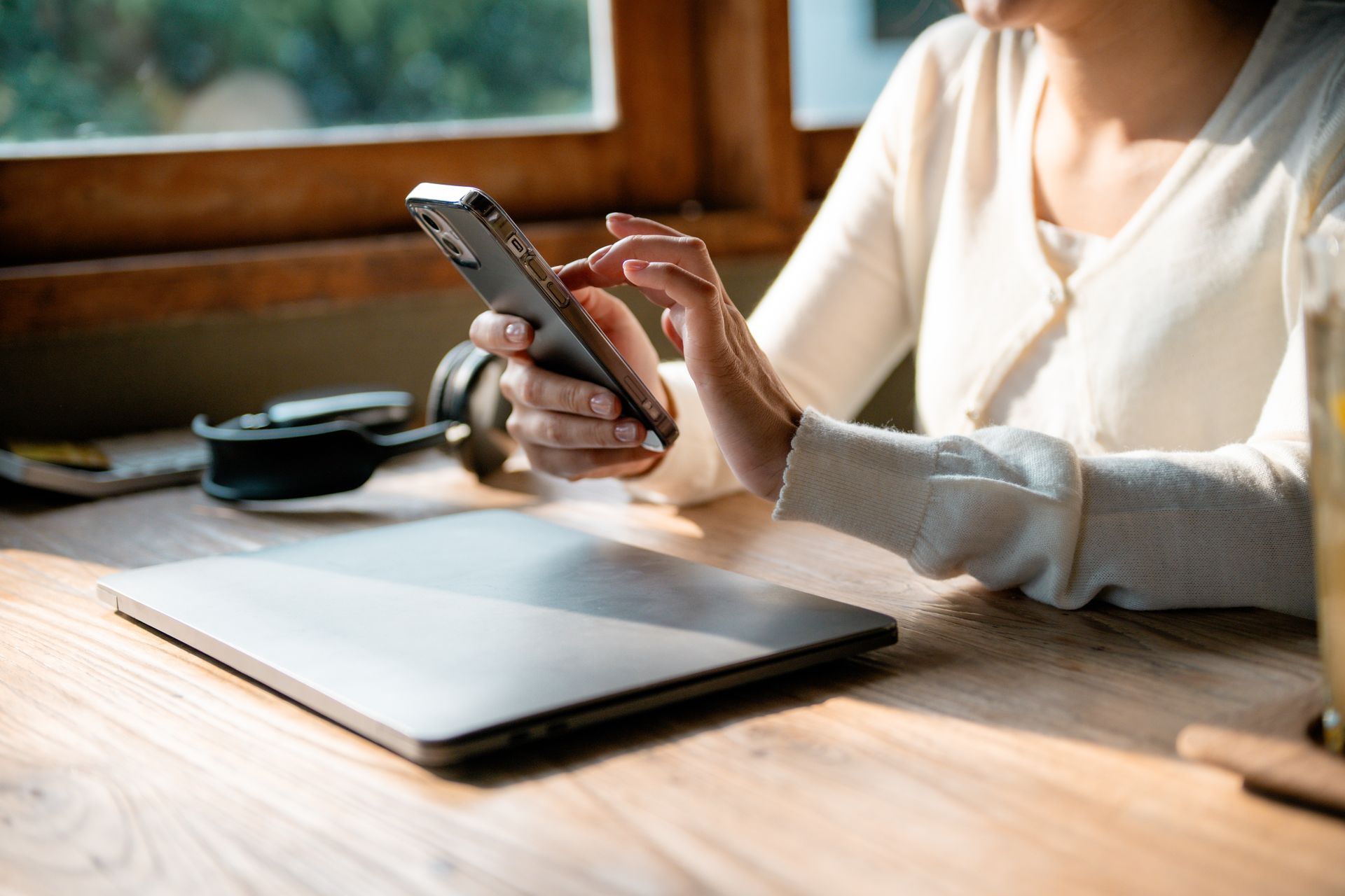 Person using a smartphone beside a closed laptop on a sunlit wooden table