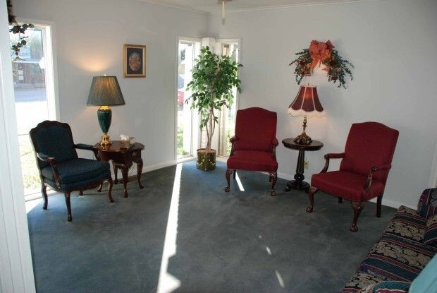 Small living room with blue carpet, two red chairs, black chair, lamp, tables, and potted plants.