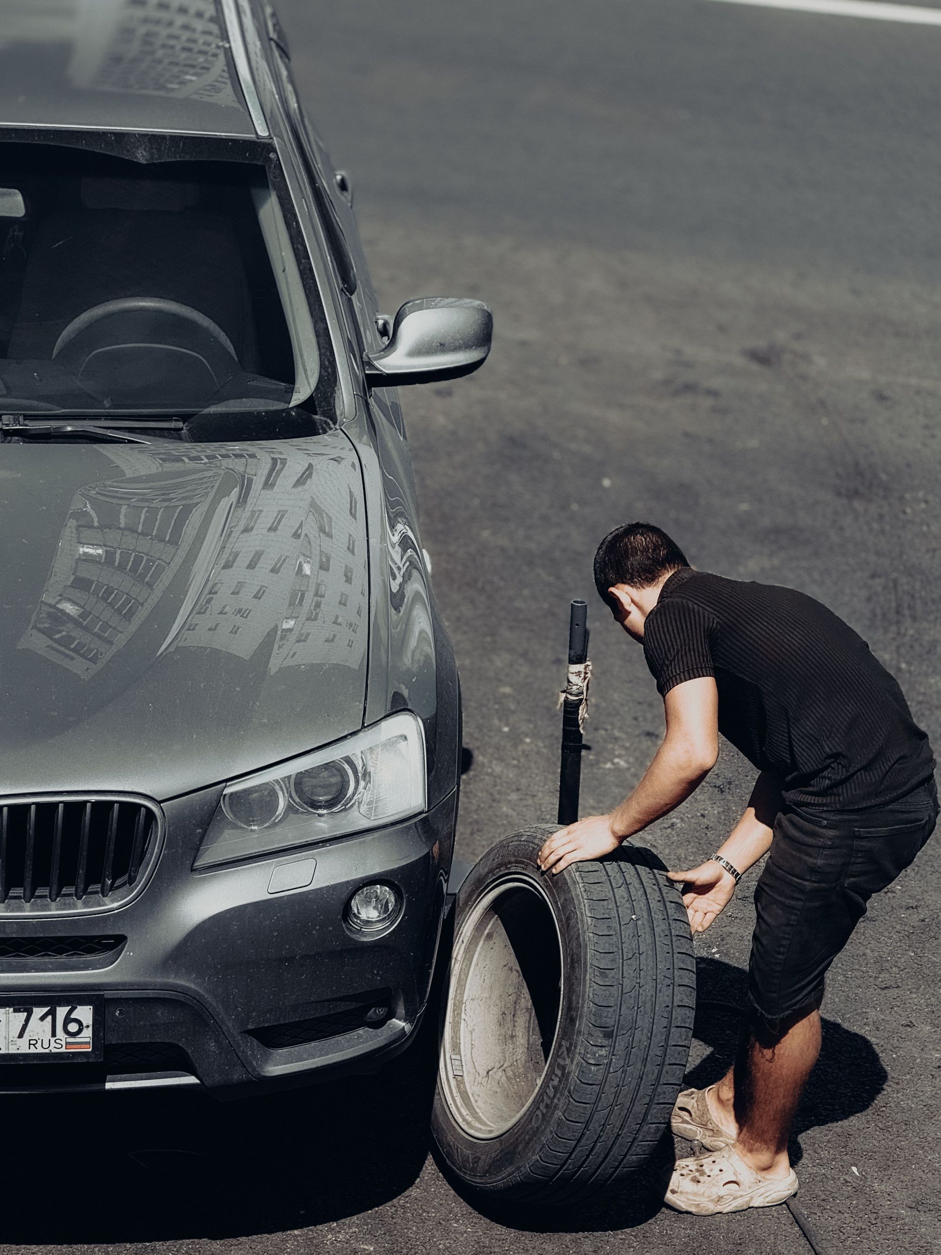 Man changing a tire beside a gray car on a roadside, holding the wheel upright.