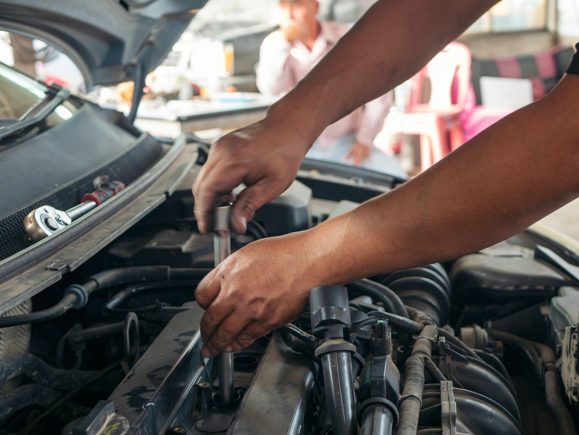 Hands working under a car hood, inspecting engine components in a garage.