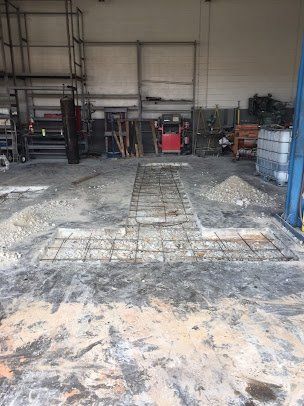 Warehouse interior with a damaged concrete floor and exposed rebar repair area near shelving and barrels