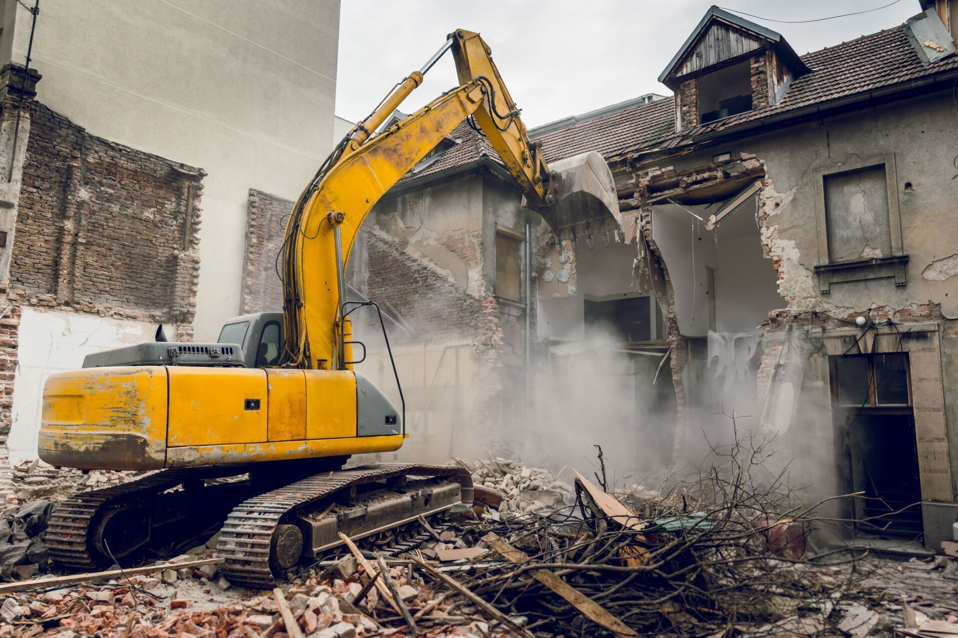Yellow excavator demolishing a damaged building, kicking up dust and rubble