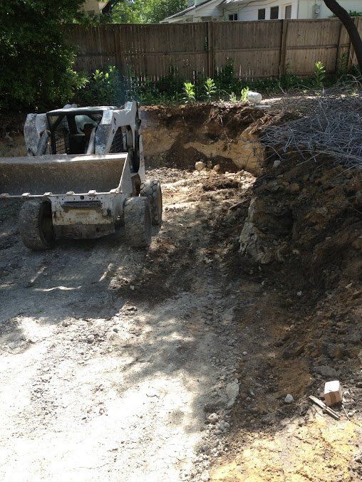 Skid steer loader beside a large dirt excavation in a backyard, with a wooden fence in the background.